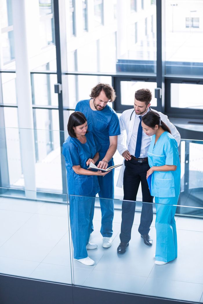 Medical team in blue scrubs reviewing a clipboard near windows. One man wears a white coat.