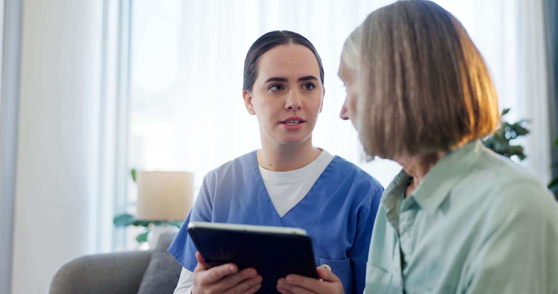 A healthcare worker in blue scrubs shows a tablet to an older woman in a home setting, discussing information.