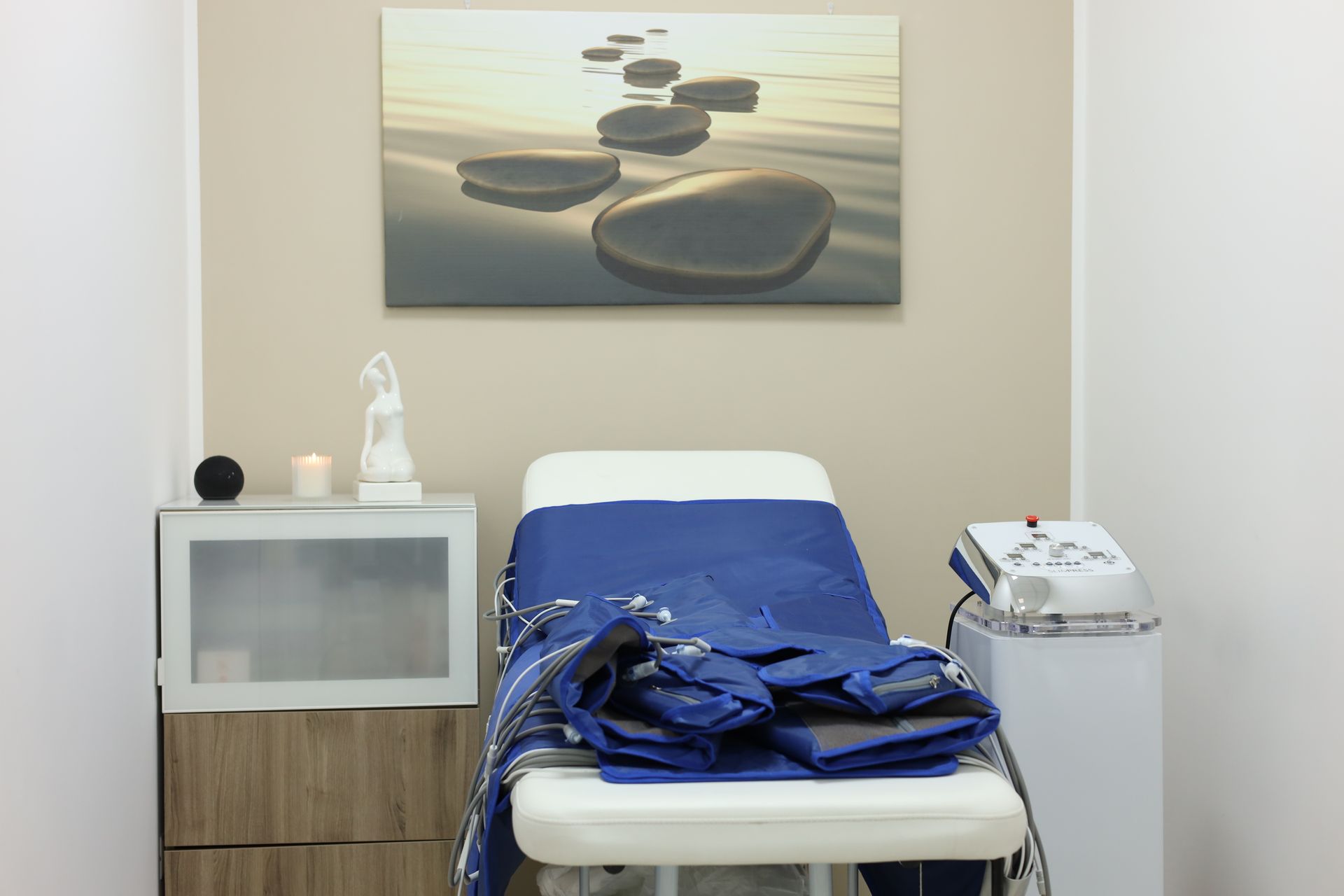 Stanza con lettino per massaggi, attrezzatura per pressoterapia e un'immagine di pietre su una spiaggia.