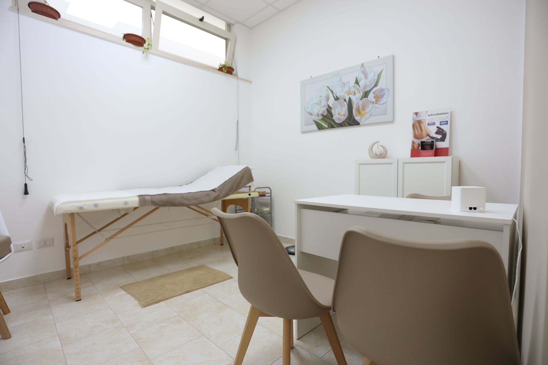 Sala visite con scrivania bianca, lettino medico, due sedie e un piccolo poster decorativo.