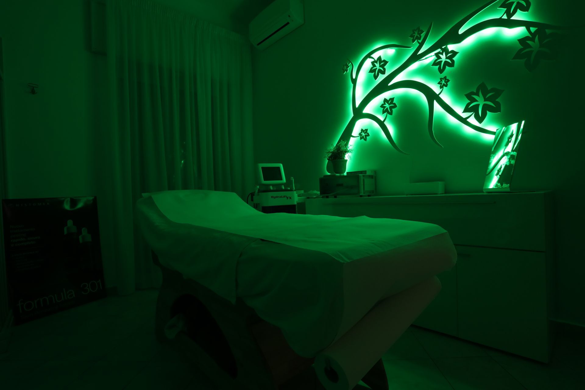 Sala benessere illuminata di verde con lettino per massaggi, attrezzature mediche e una parete luminosa raffigurante una pianta.