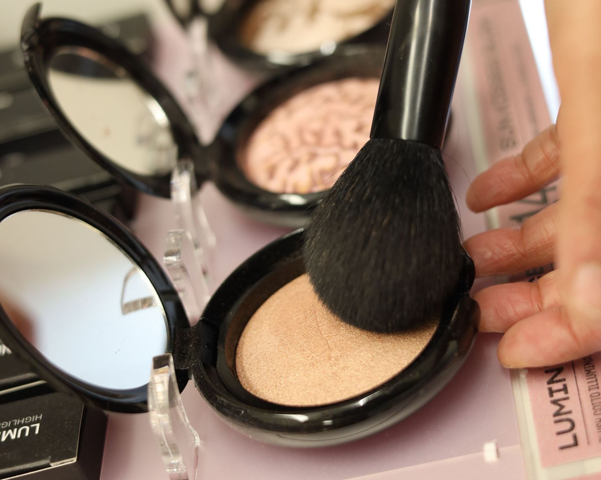 Mano con pennello da trucco che applica l'illuminante dal cofanetto aperto.