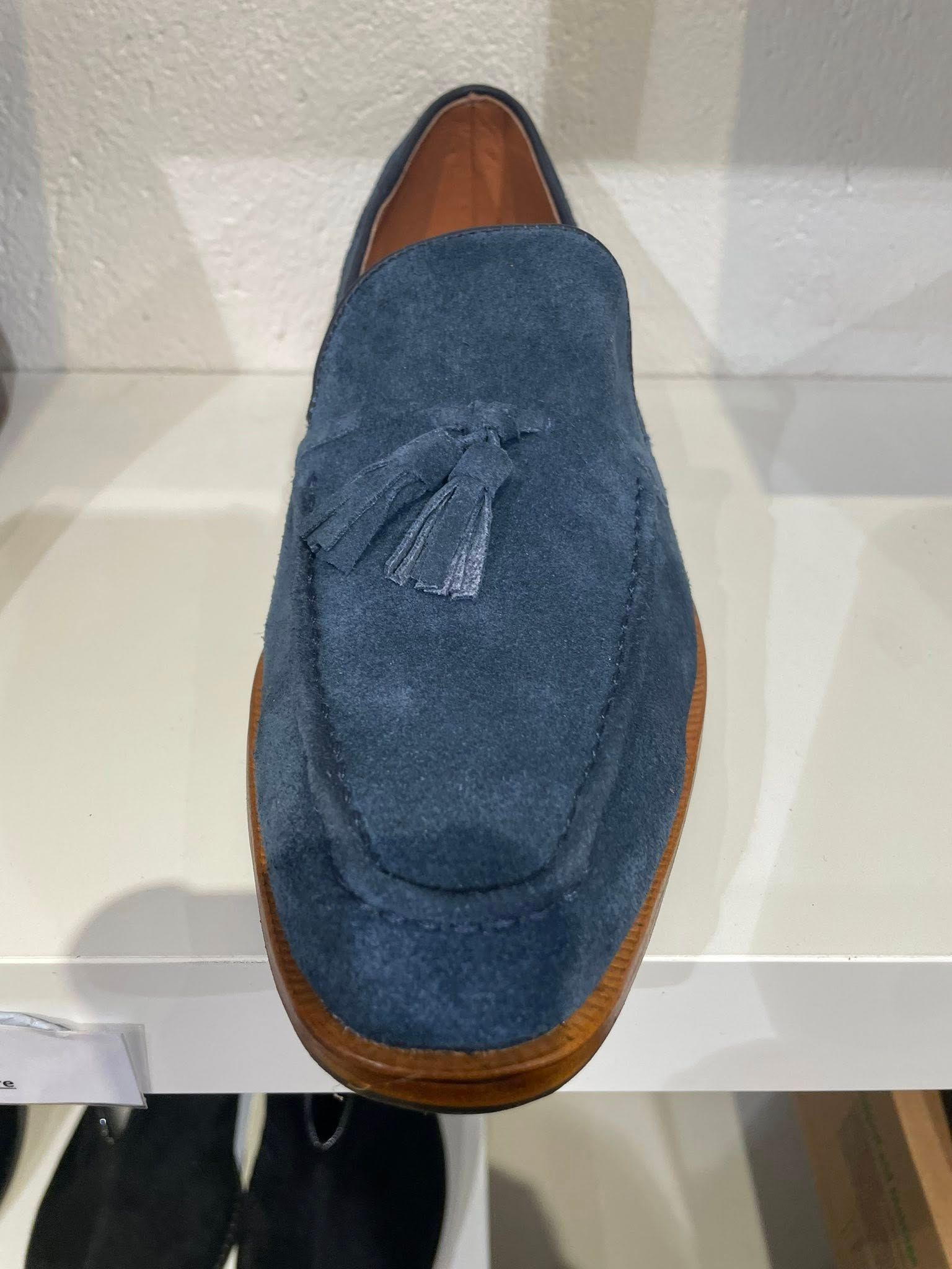 Blue suede loafer with tassel, on a white shelf.