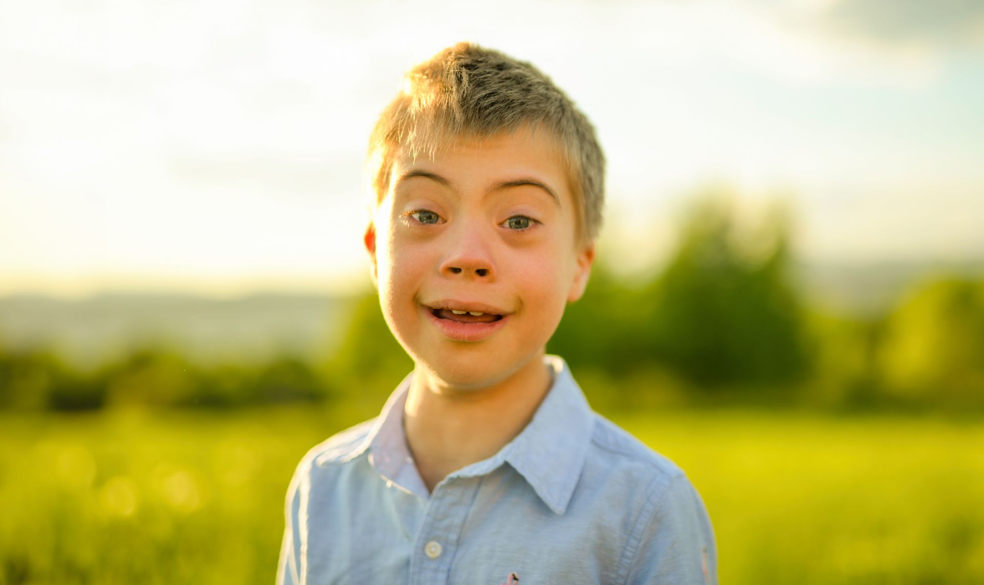A young boy with down syndrome is standing in a field and smiling.
