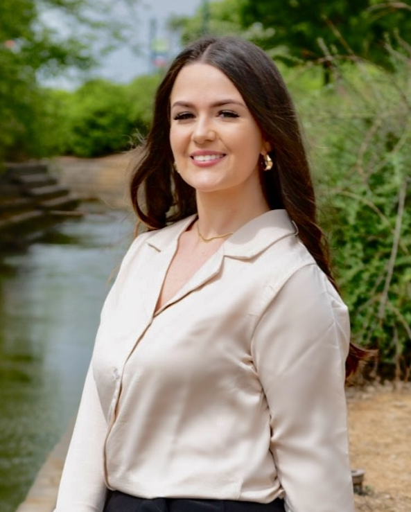 Woman in cream-colored blouse smiles near a body of water in a natural setting.