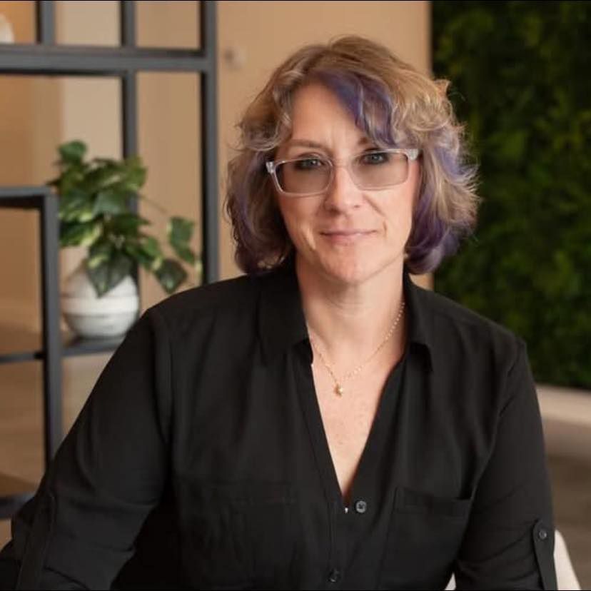 Woman in black shirt and glasses smiles, short hair with purple highlights, in office setting.