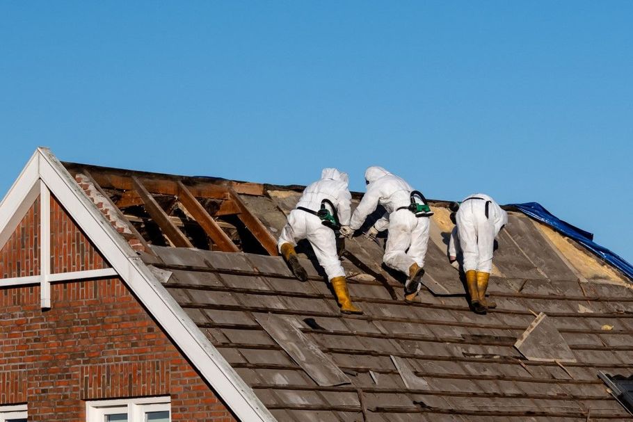 Three workers in white suits repair a damaged roof under a blue sky.