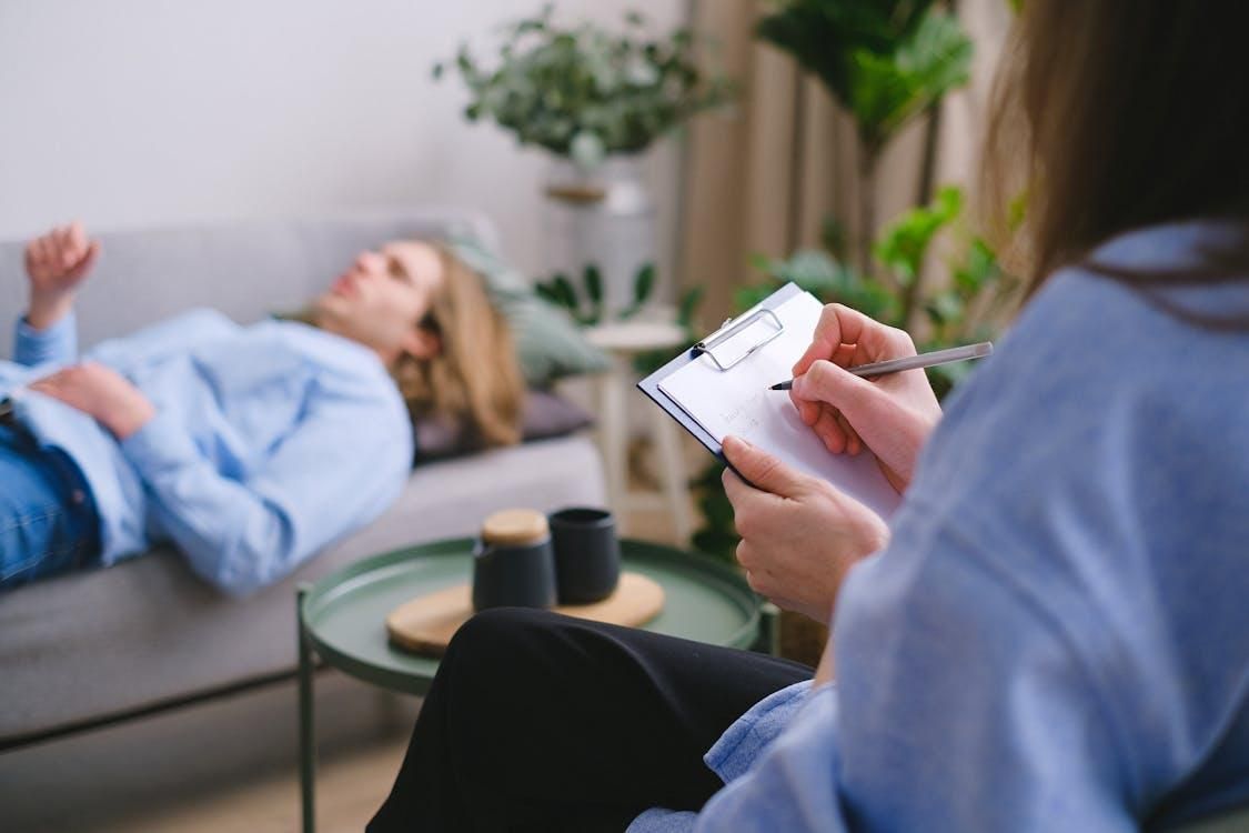 A cropped image showing a psychotherapist holding a clipboard and writing notes with a patient lying