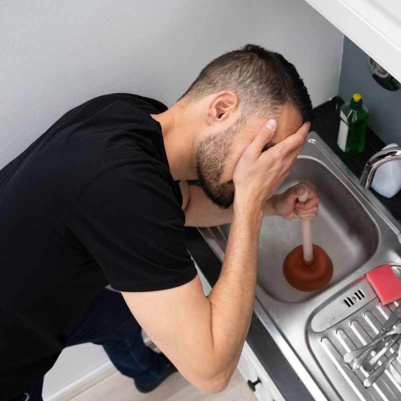 Man using plunger in a kitchen sink, looking frustrated, hands covering face.
