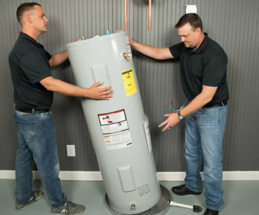 Two people lift and maneuver a cylindrical water heater in a gray-walled room.