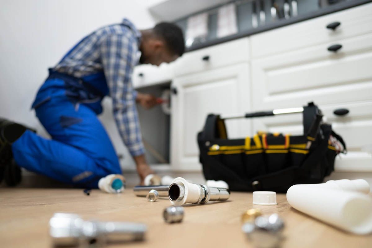 Plumber working under a kitchen sink, with tools and plumbing parts on the floor.