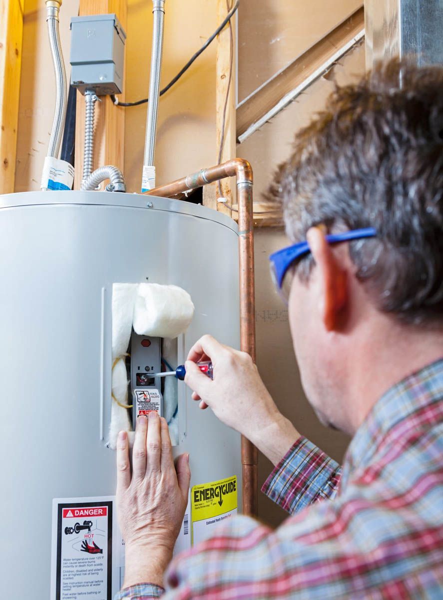 Person fixing a water heater; using a screwdriver. Wearing safety glasses. Indoors, near wood paneling.