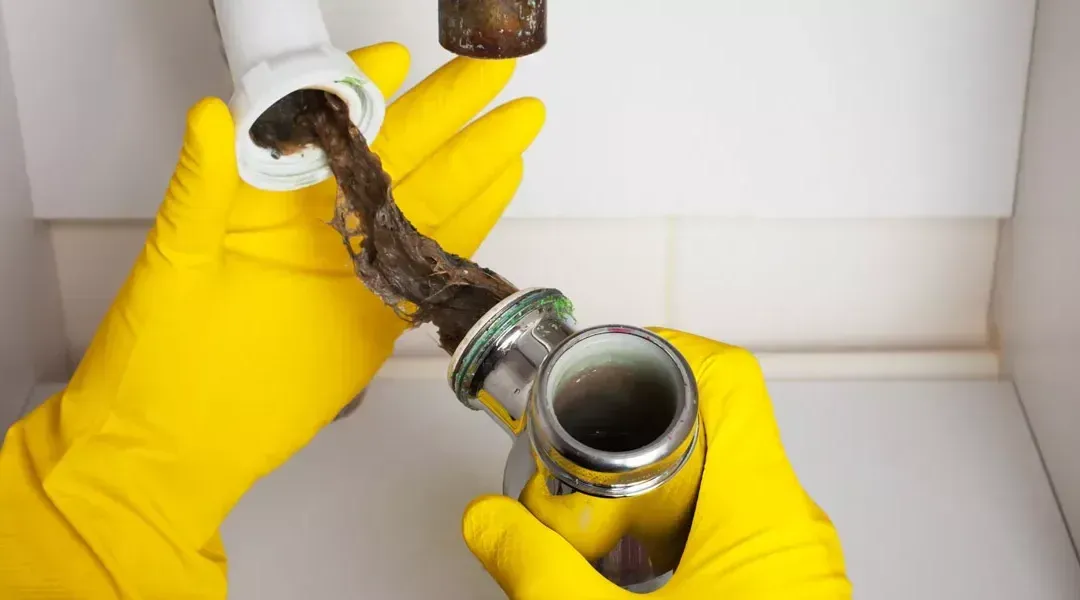 Hands in yellow gloves removing a clogged drain pipe from a sink.