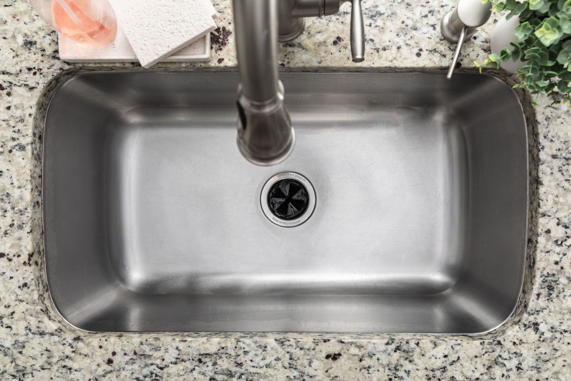 Stainless steel kitchen sink in granite countertop, viewed from above. Faucet centered above drain.
