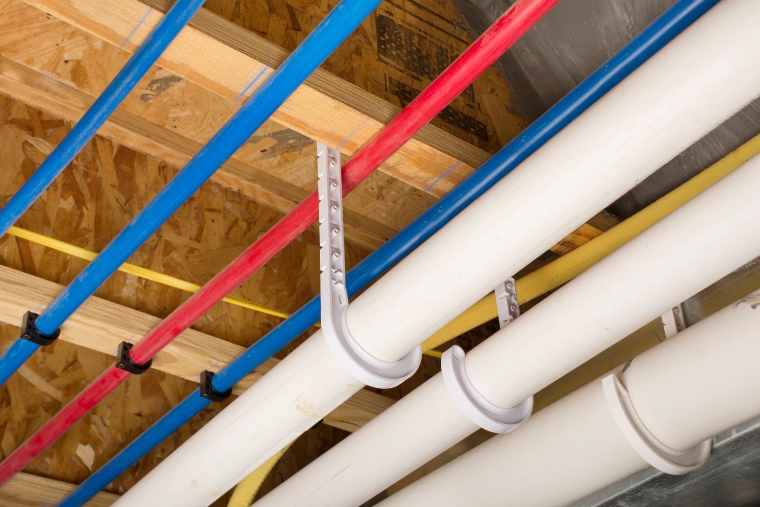 Blue, red, yellow, and white pipes secured to wooden ceiling joists, likely for water and drainage.