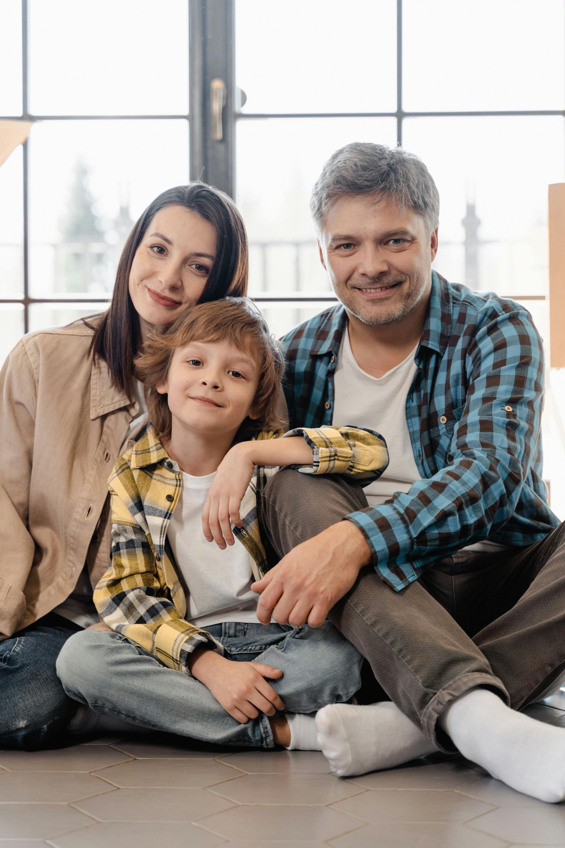 Family of three smiling, sitting on a floor by a window. The boy is wearing a yellow plaid shirt.