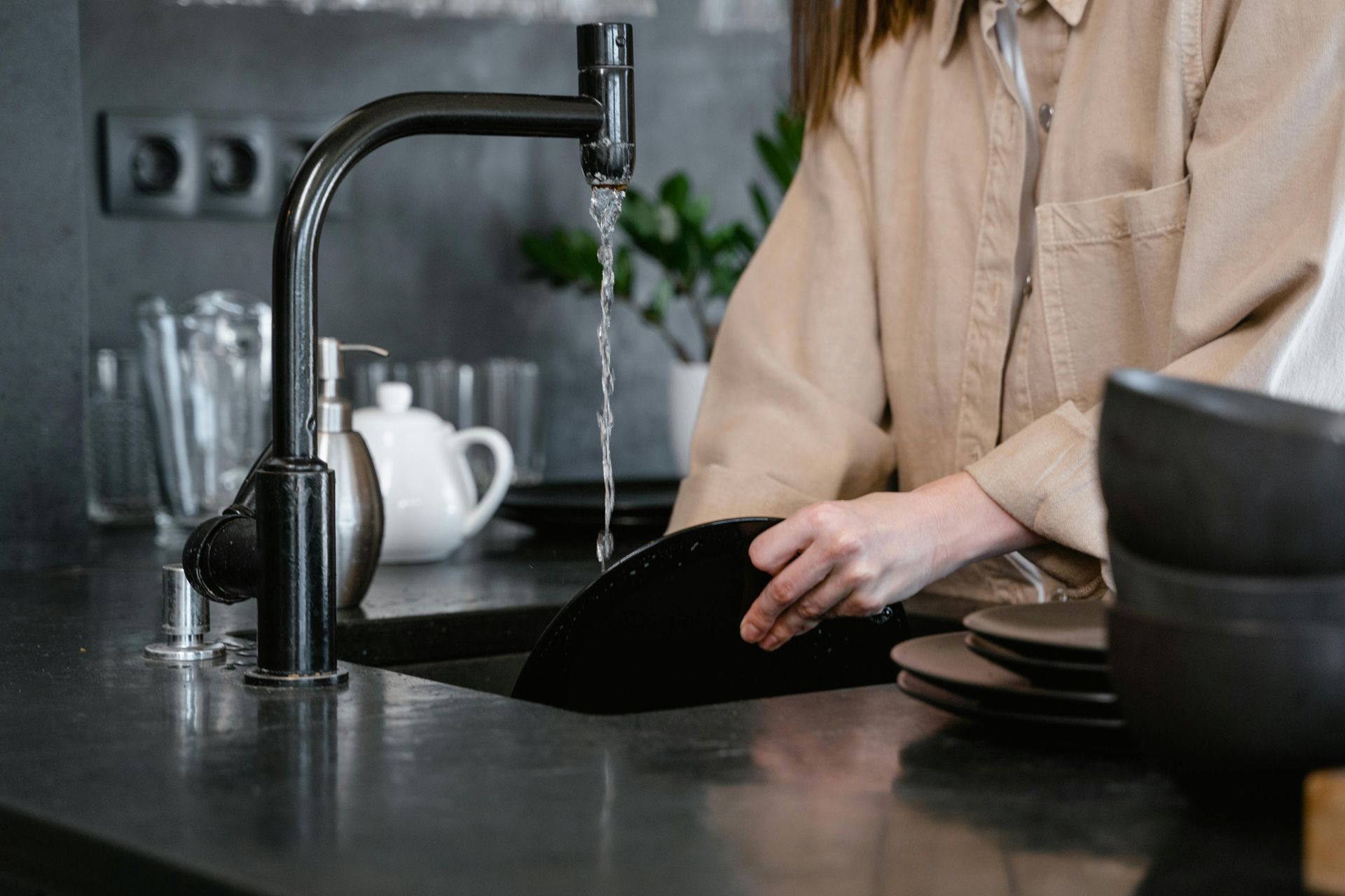 Person washing a black plate in a black kitchen sink under a running faucet.