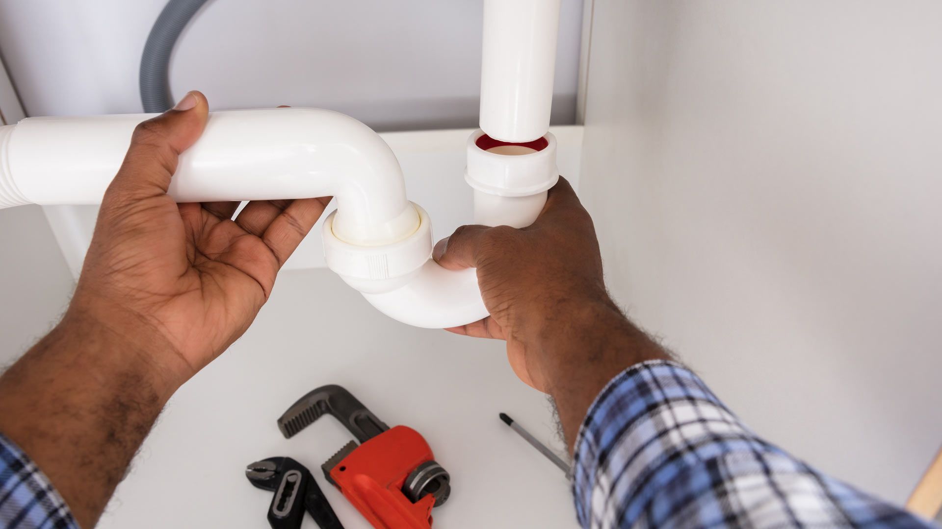 Hands connecting white plumbing pipes under a sink. A wrench and screwdriver are in the foreground.