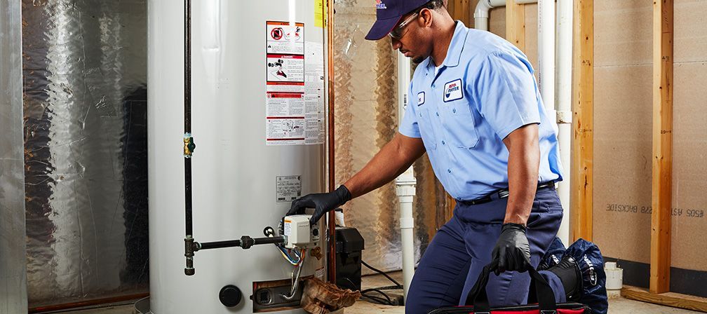 Plumber in blue uniform examines a water heater in a basement, holding a tool bag.