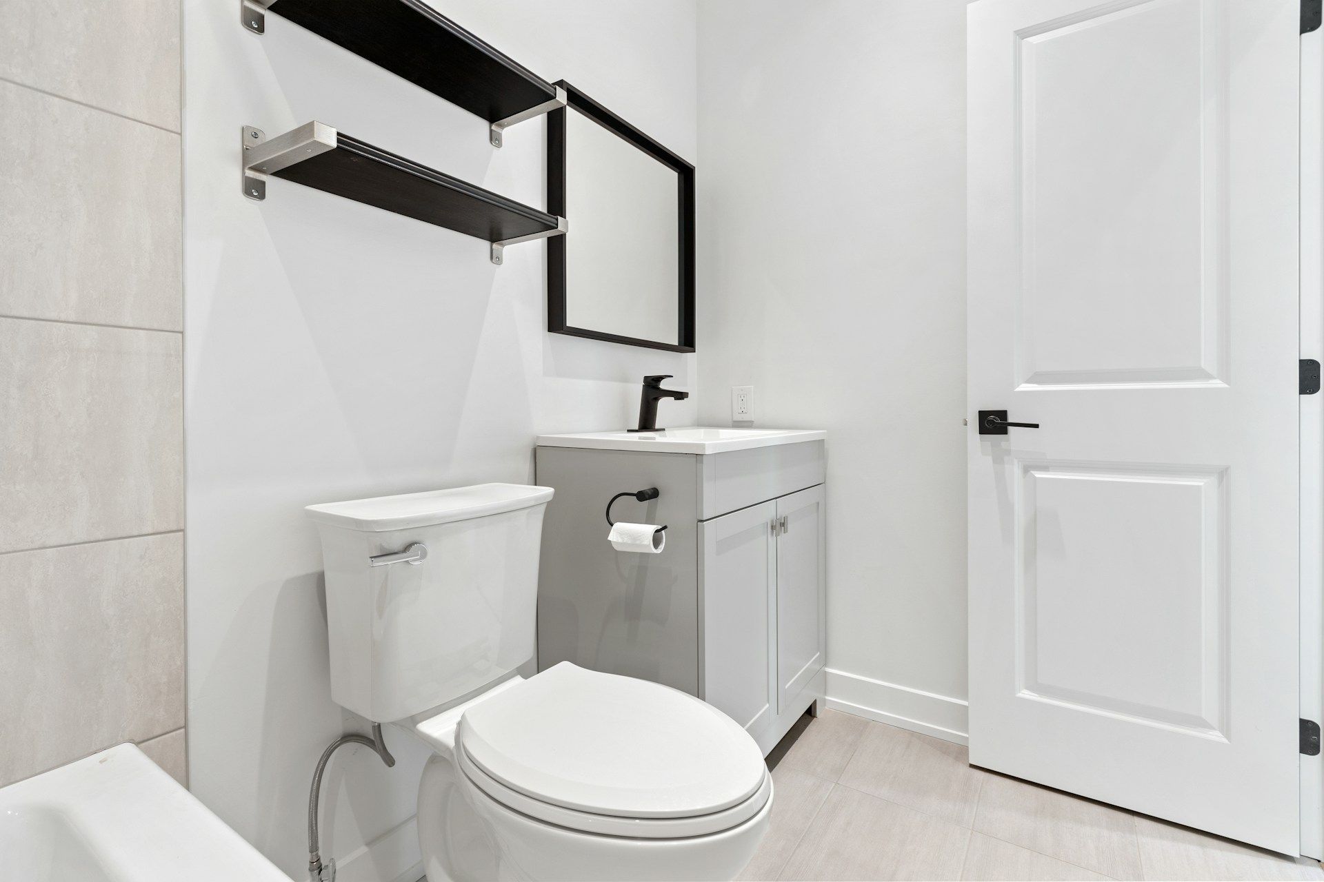 Bathroom with white walls, toilet, vanity, black-framed mirror, shelves, and a closed door.