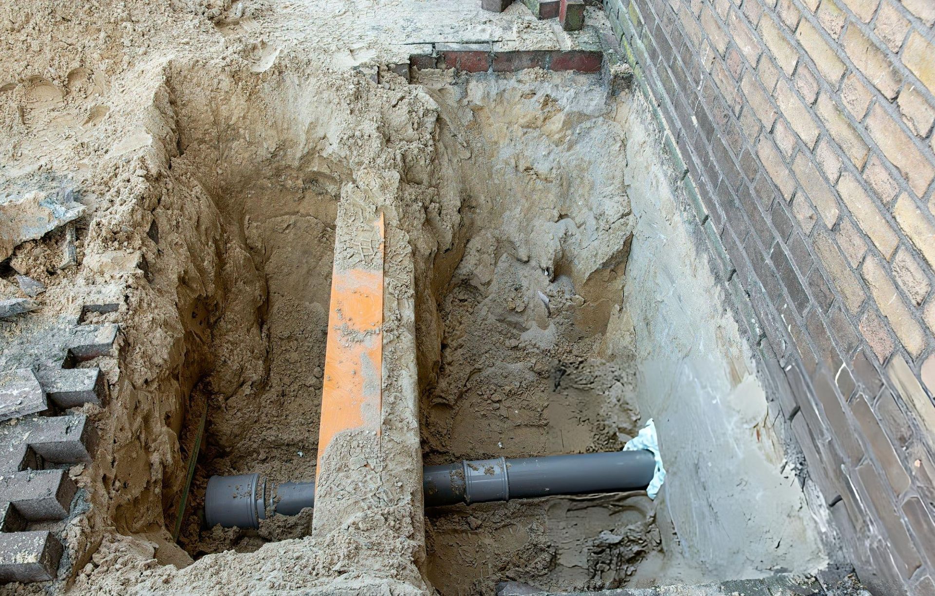 Excavation revealing buried plumbing pipe and orange marker near brick wall.