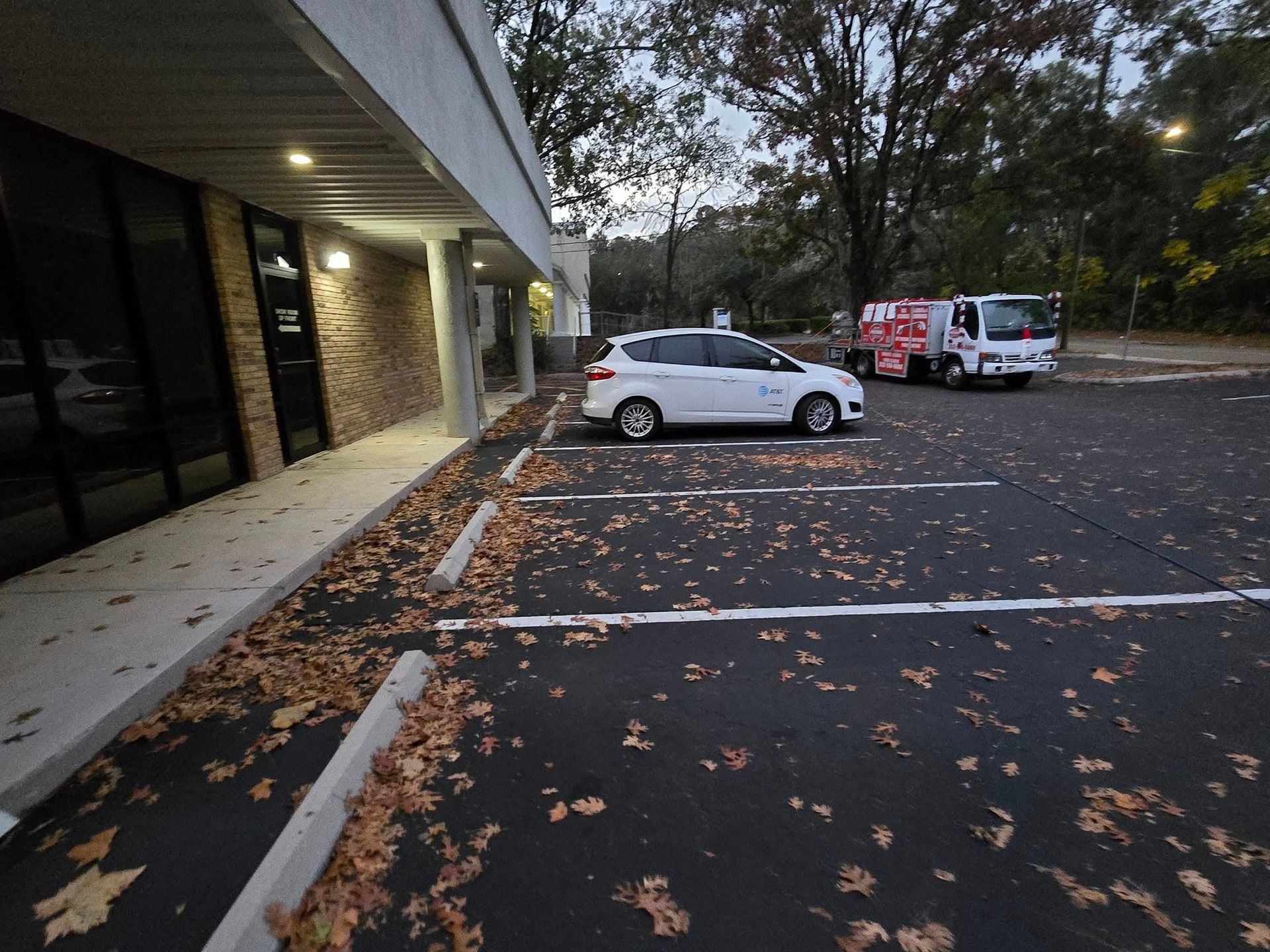 White SUV parked near a commercial building and a service truck in a parking lot covered with leaves.