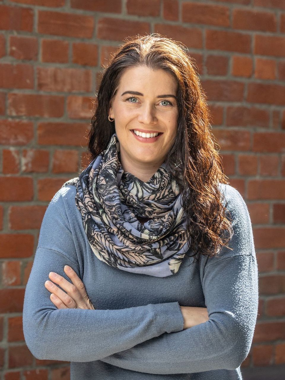 A woman wearing a scarf and a blue sweater is standing in front of a brick wall.