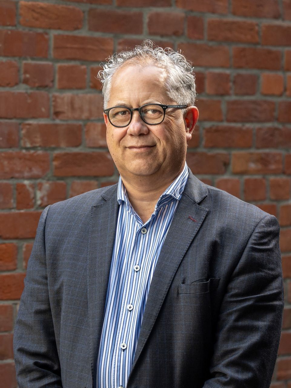 A man in a suit and glasses is standing in front of a brick wall.