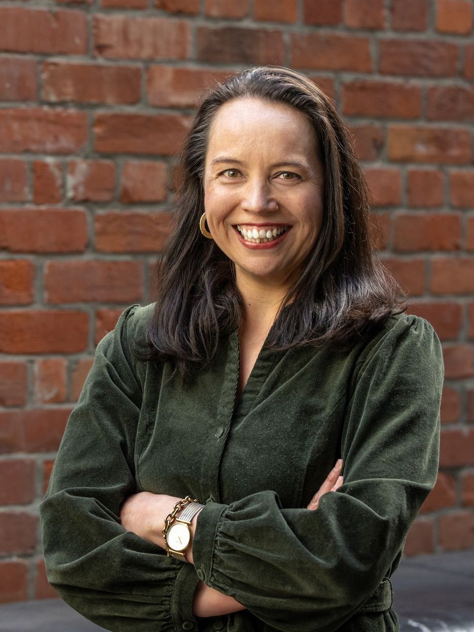 A woman in a green shirt is standing in front of a brick wall with her arms crossed and smiling.