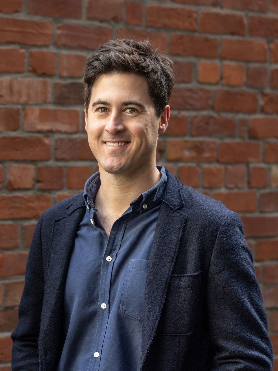 A man in a suit and blue shirt is standing in front of a brick wall.