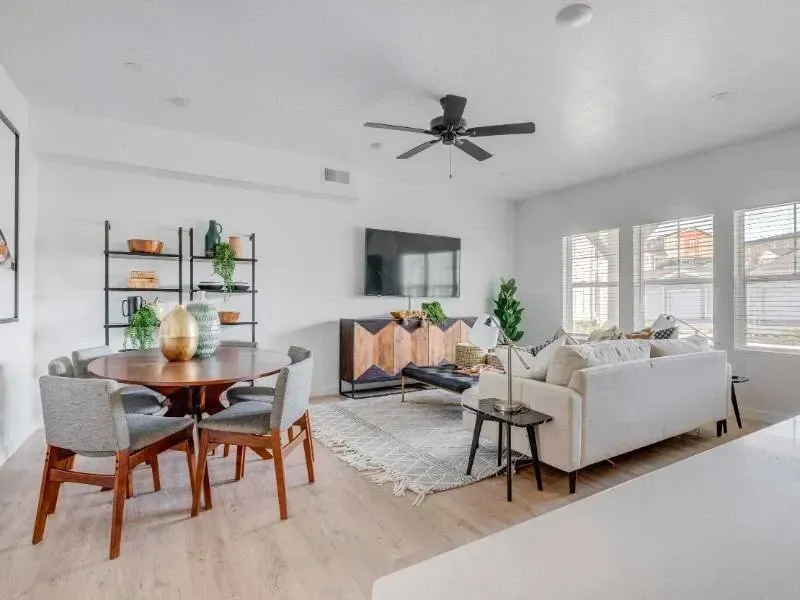 Bright living room with a beige sofa, round dining table, TV, open shelving, and large windows.