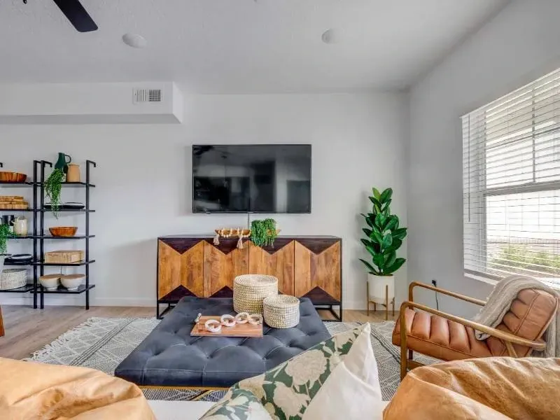 Bright apartment living room with a wall-mounted TV, geometric wood console, shelving, and a large window.