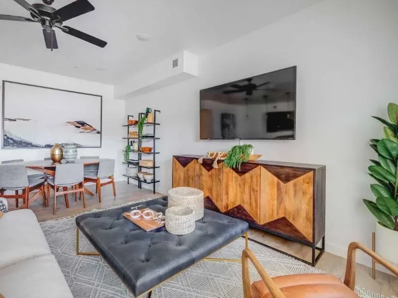 Apartment living room with a tufted ottoman, sofa, TV on wall, and dining area.