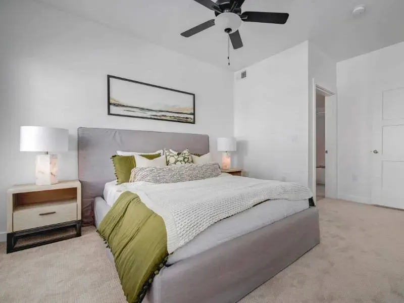Bedroom with gray upholstered bed, two wooden nightstands with lamps, and a ceiling fan.