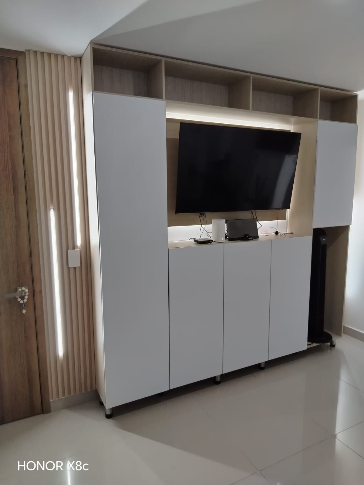 Gabinetes blancos empotrados con un televisor montado, estantes y una luz decorativa se destacan en una habitación.