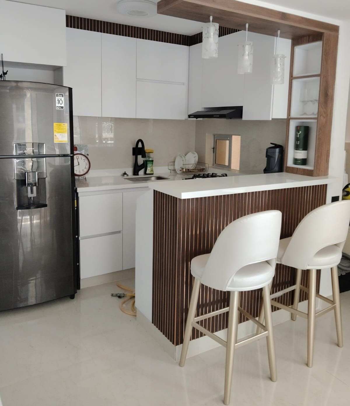 Cocina moderna blanca con refrigerador de acero inoxidable, encimeras blancas y detalles de madera. Dos taburetes de bar.