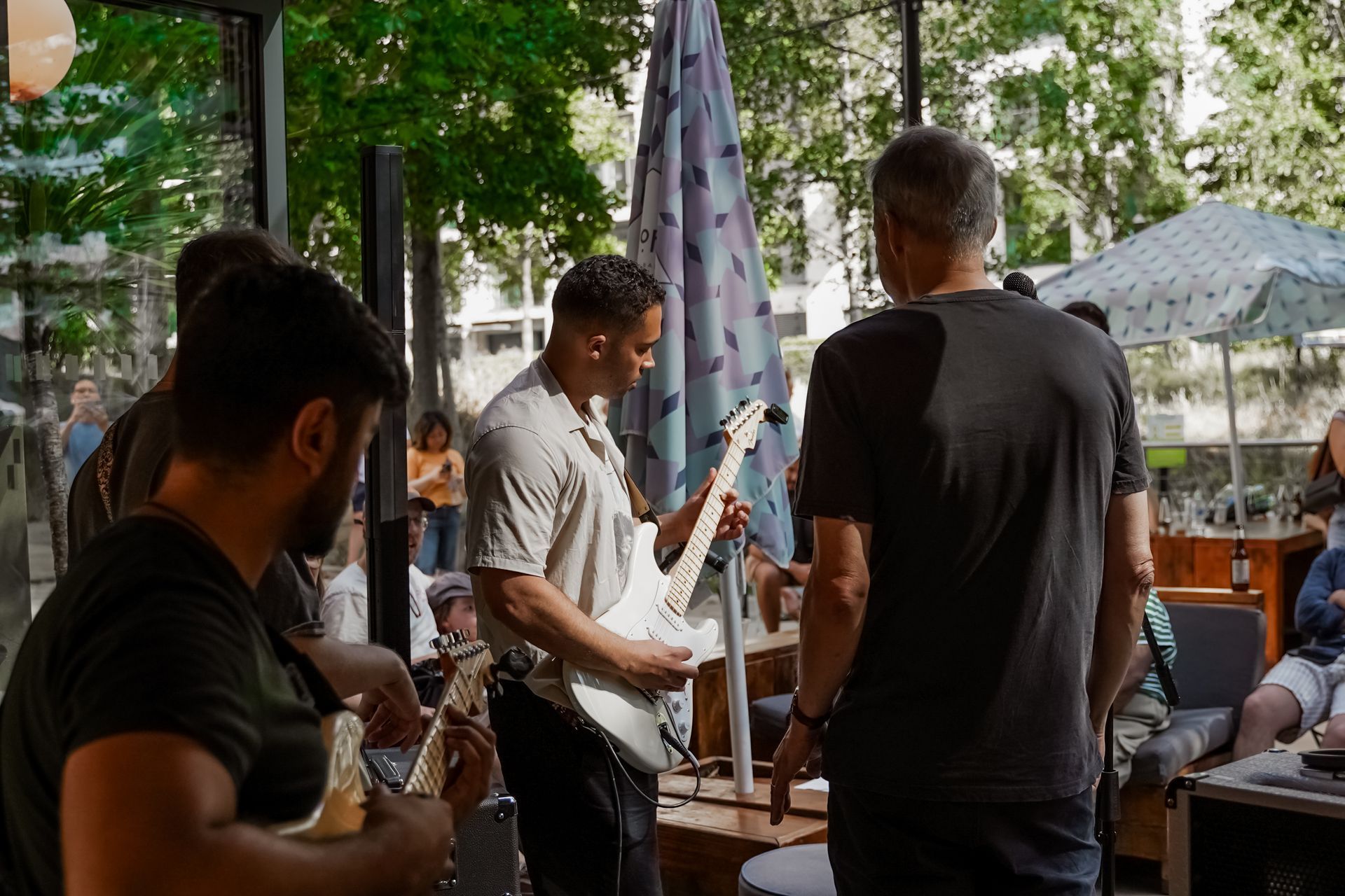 Musicians playing guitar at outdoor cafe. One man plays a white guitar. Others watch.