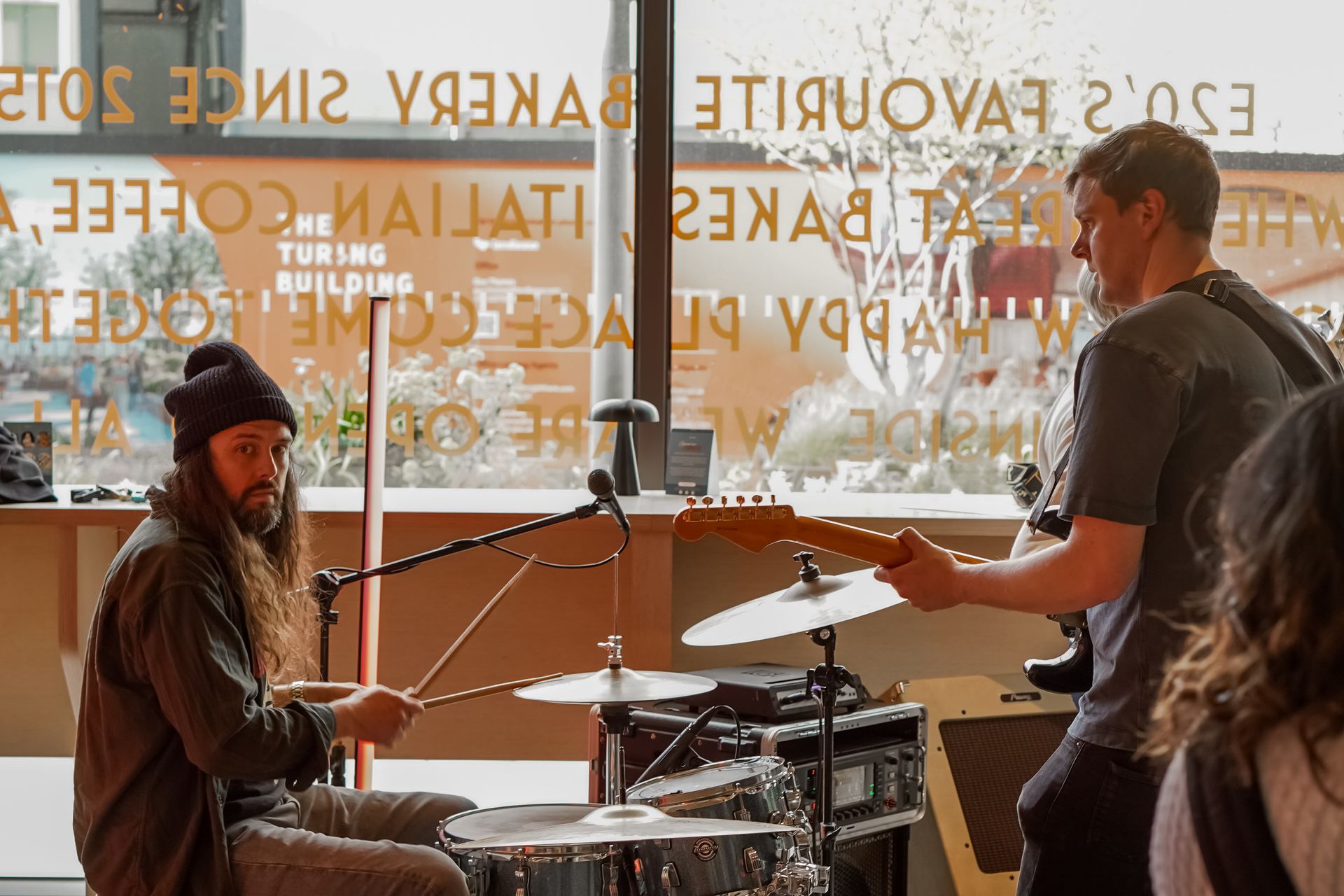 Musicians rehearsing in a cafe, with drums and guitar by a large window.