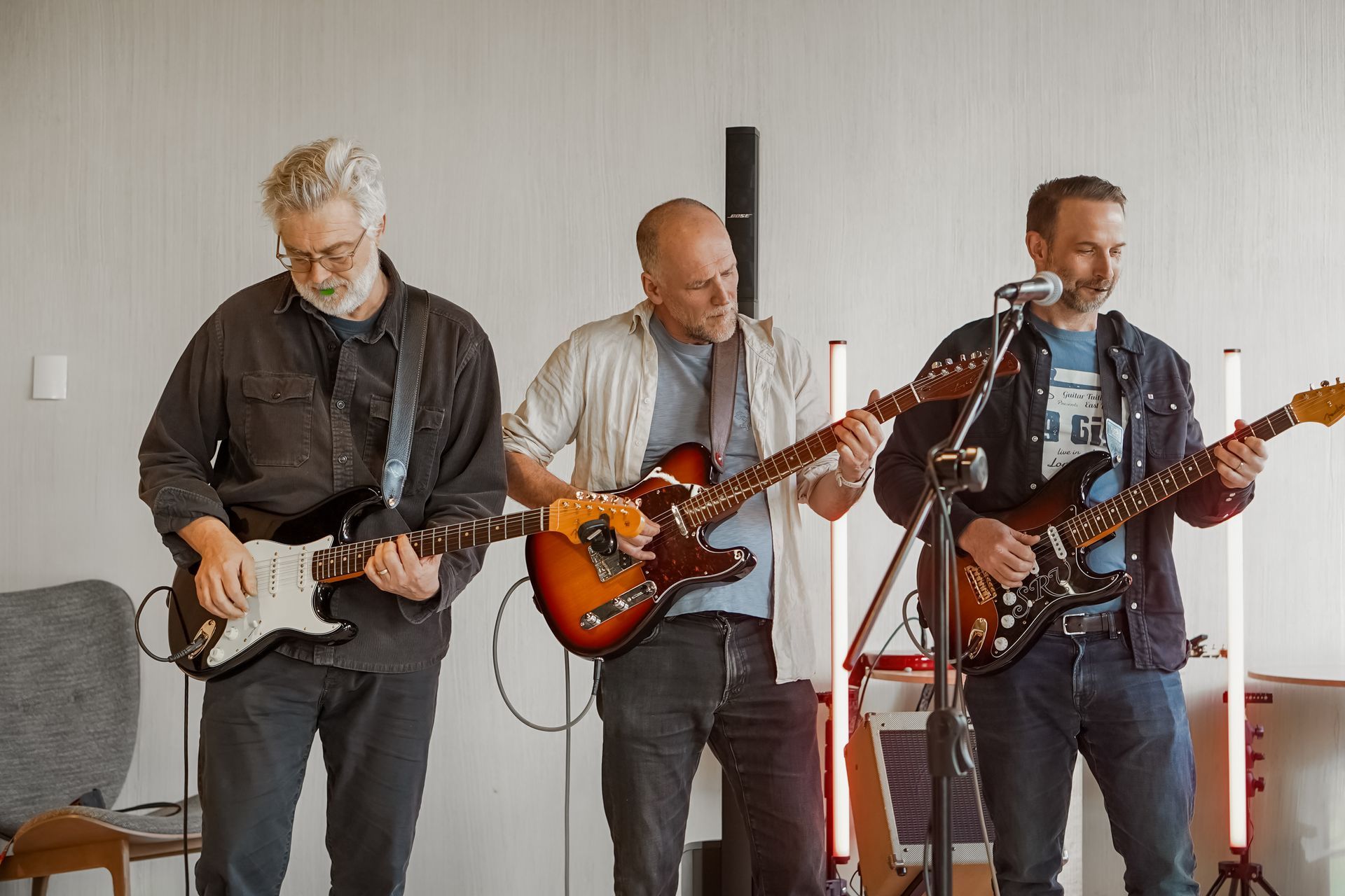 Three musicians playing electric guitars in a bright room, with a microphone stand at center.