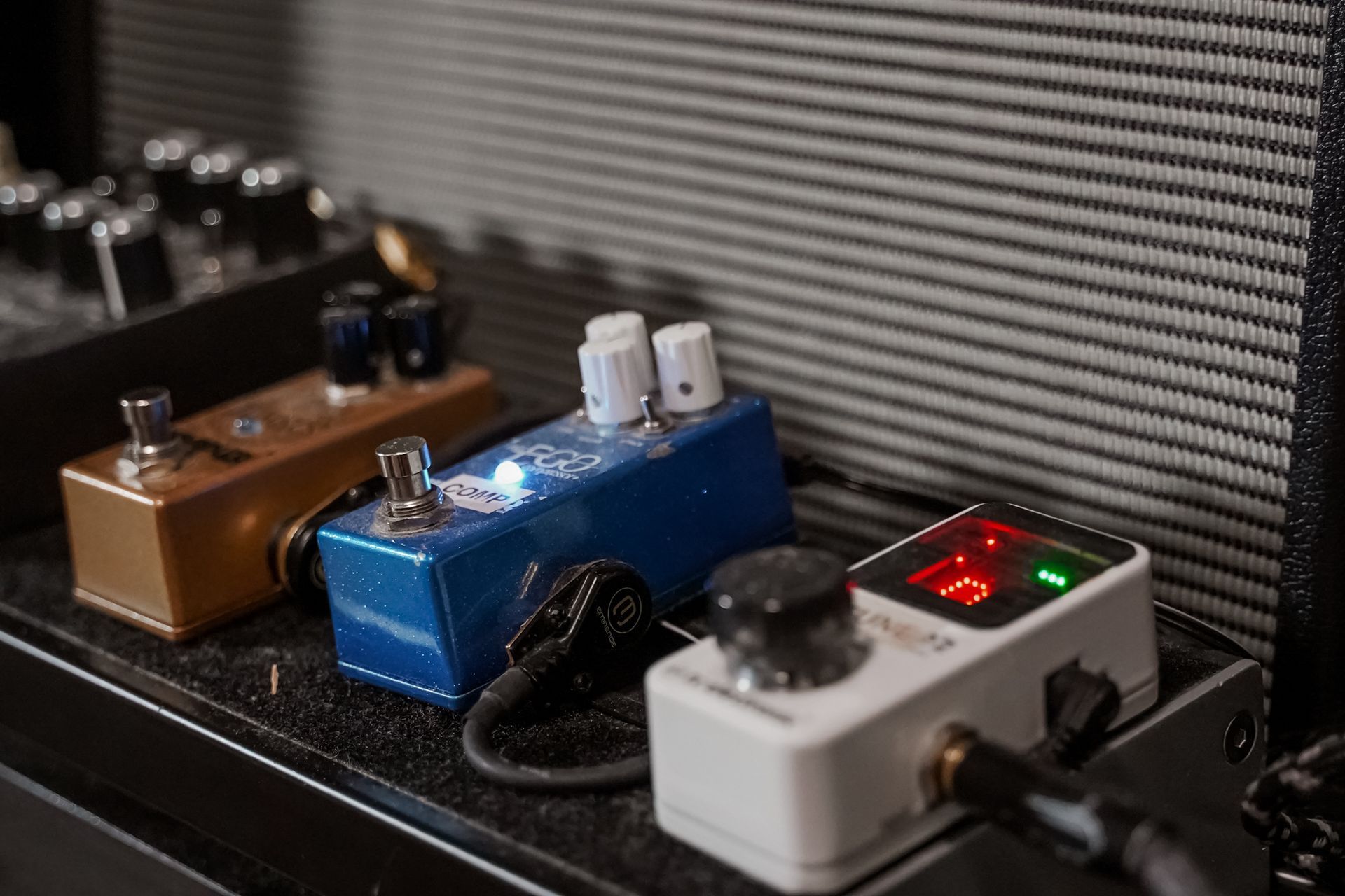 Close-up of guitar effect pedals in a row on a black surface. Gold, blue, and white pedals are visible.
