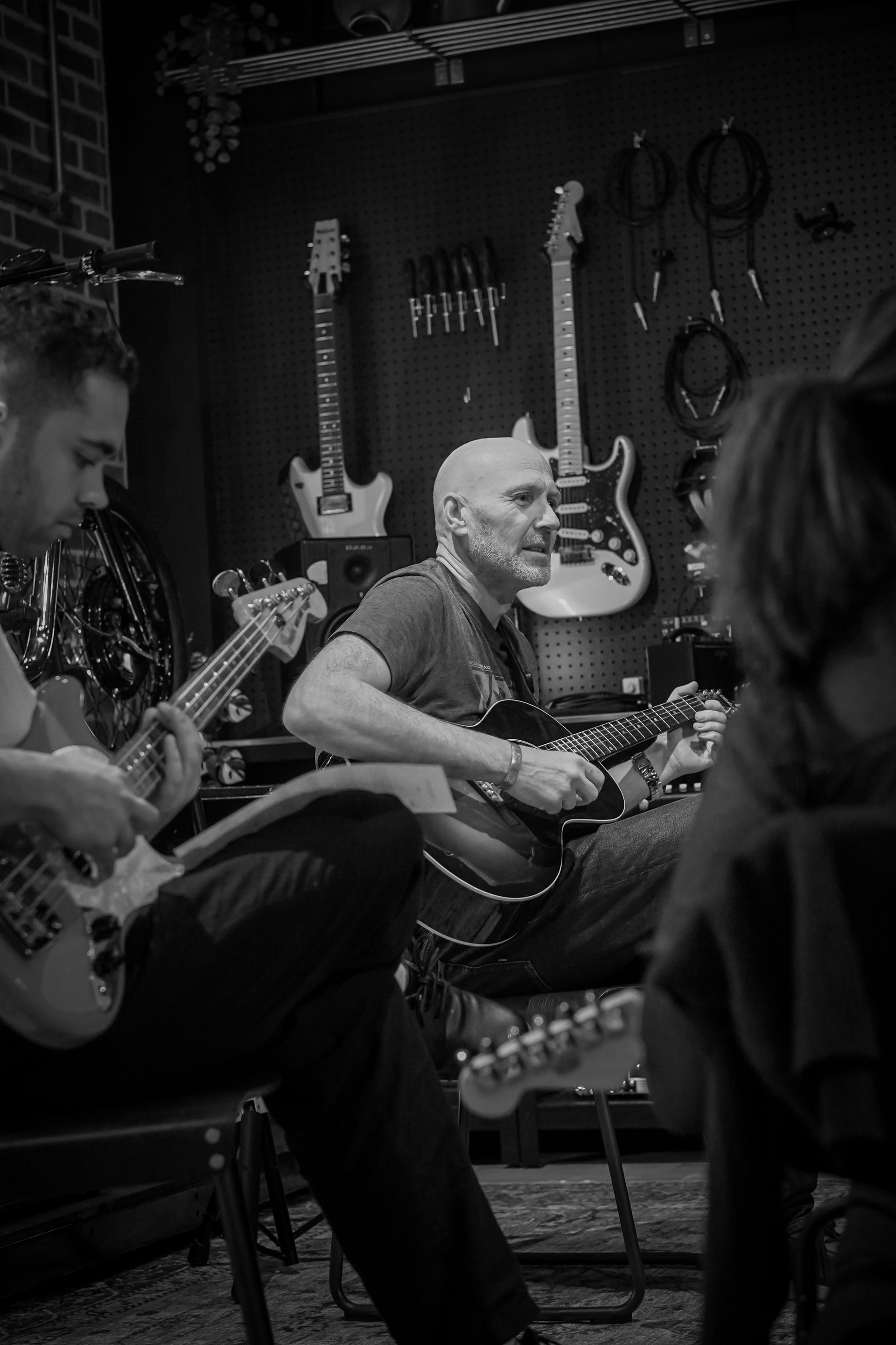 Musicians playing guitars in a music shop, with instruments hanging on the wall and an audience nearby.