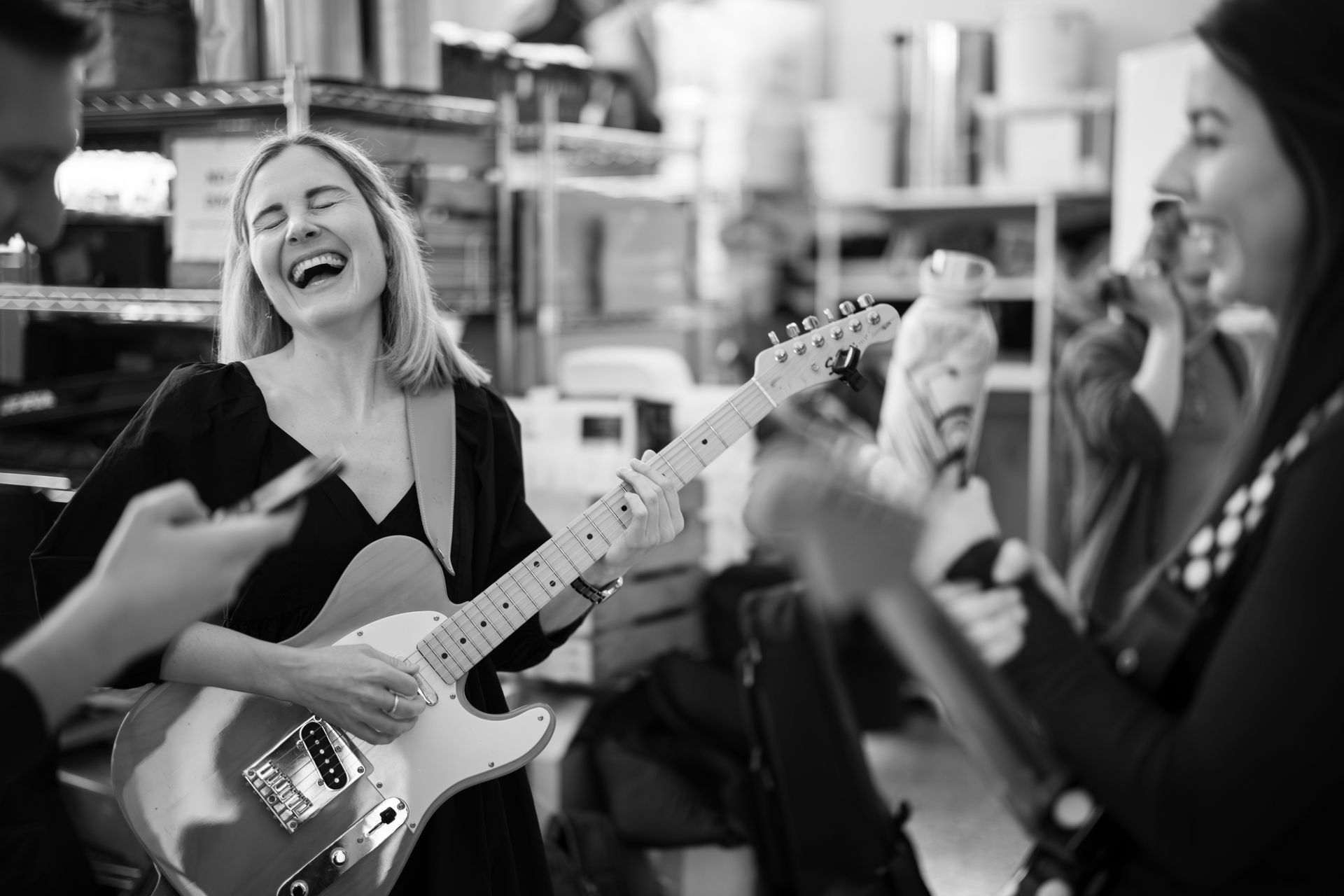Woman laughing, playing electric guitar with friends. Black and white photo, indoor setting.
