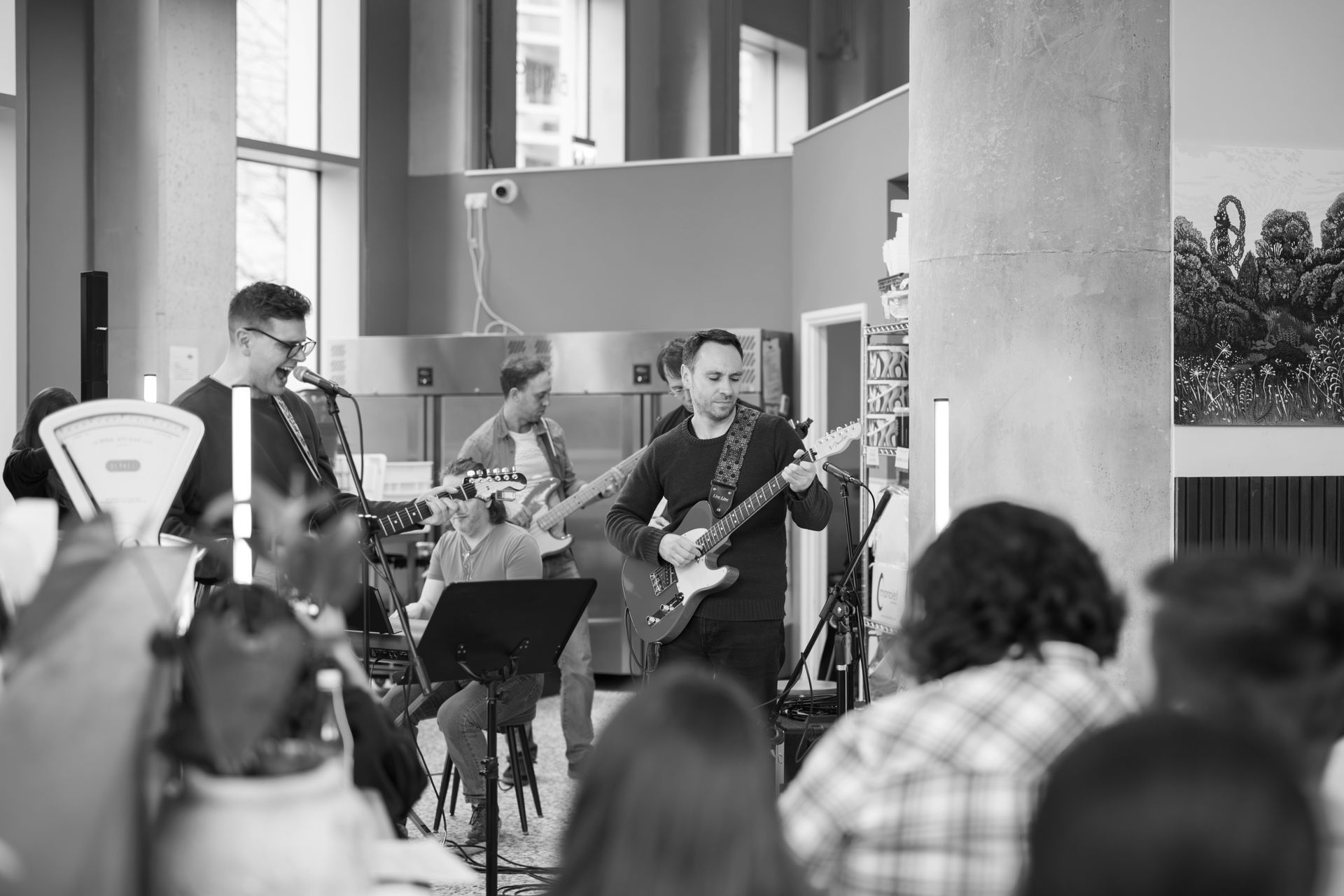 Band playing music indoors, with people in the audience. Instruments include a guitar and microphone.