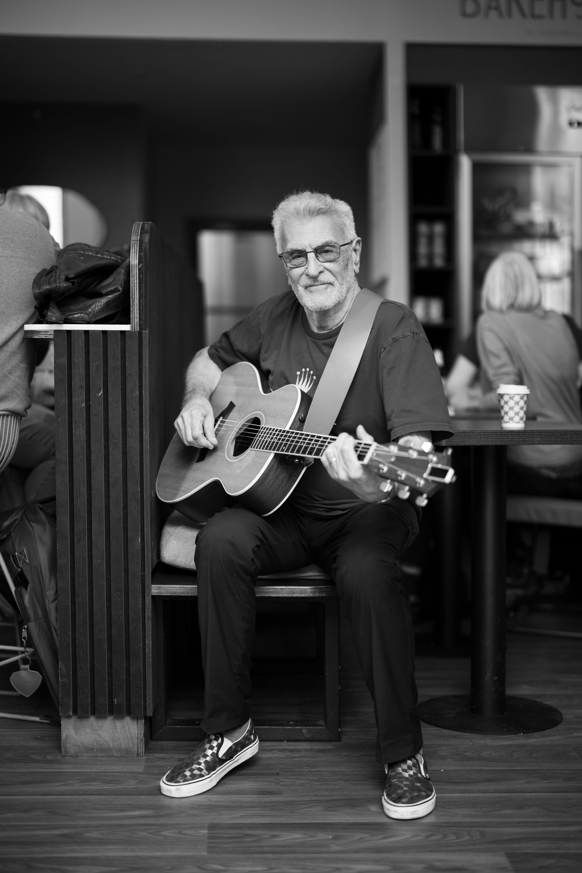 Older man with gray hair and sunglasses playing acoustic guitar in a cafe.