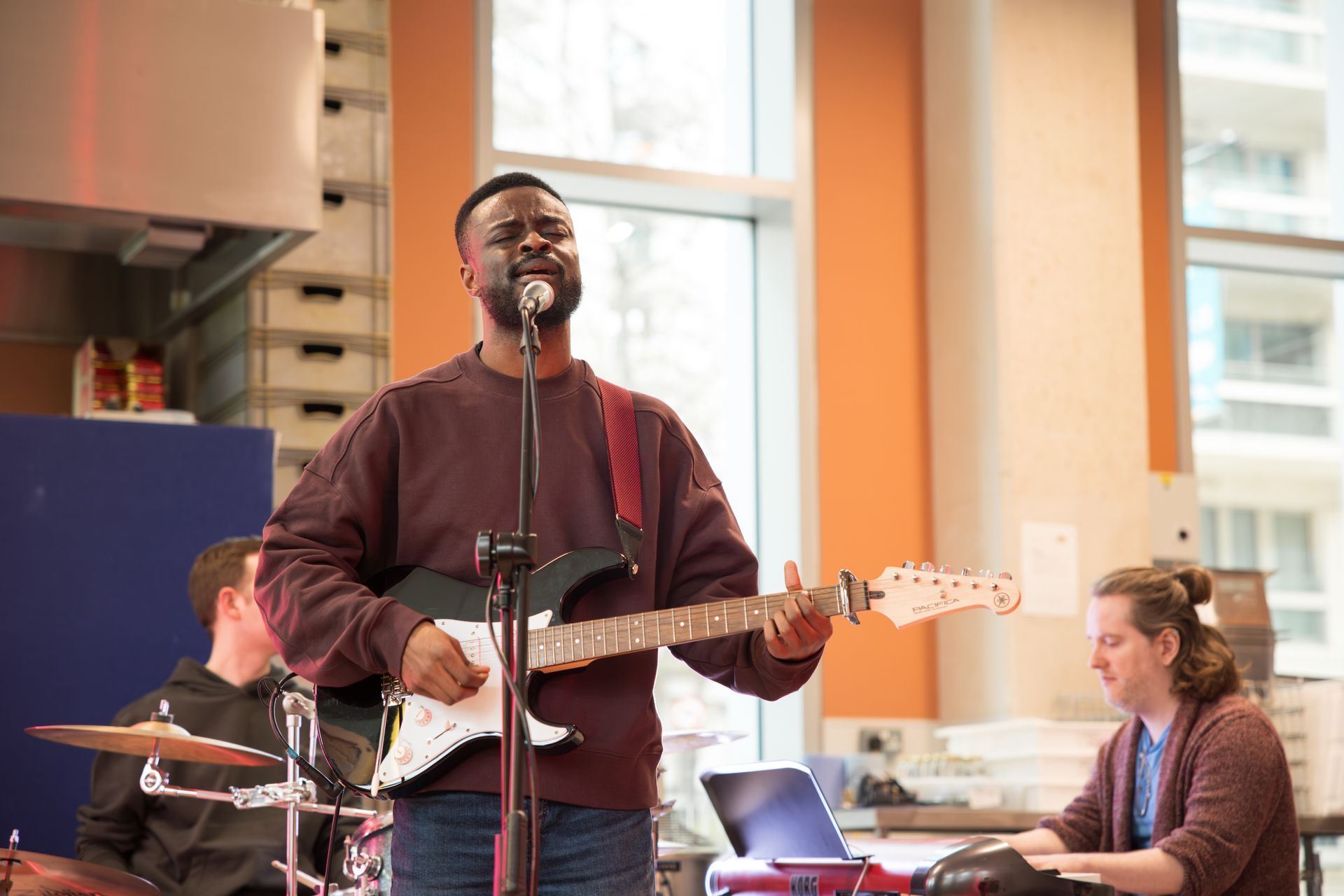 Man playing guitar and singing into a microphone, with a drummer and keyboard player in the background.