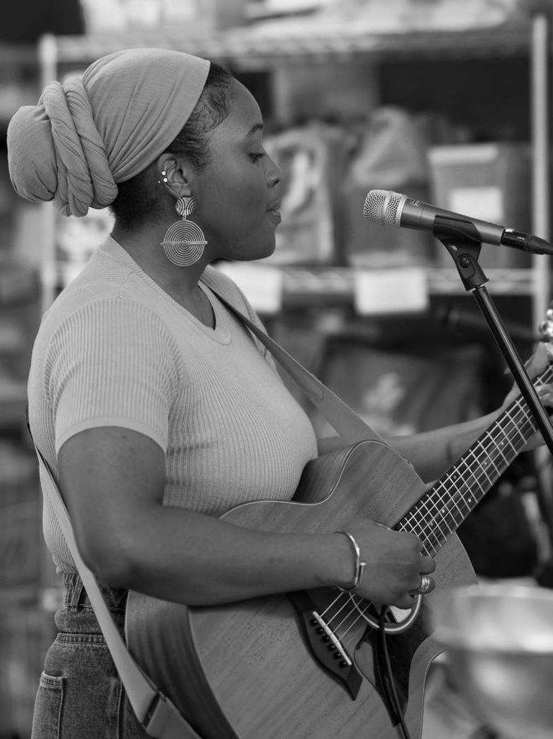 Black and white photo: Woman with headscarf sings while playing a guitar at a venue, microphone in front of her.