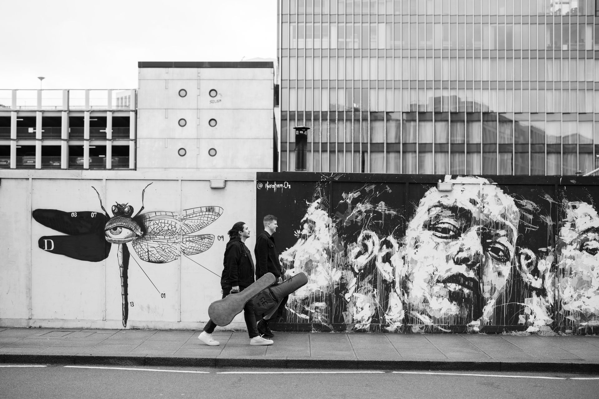 Two people walk past street art of a dragonfly and faces; grey tones, daytime.