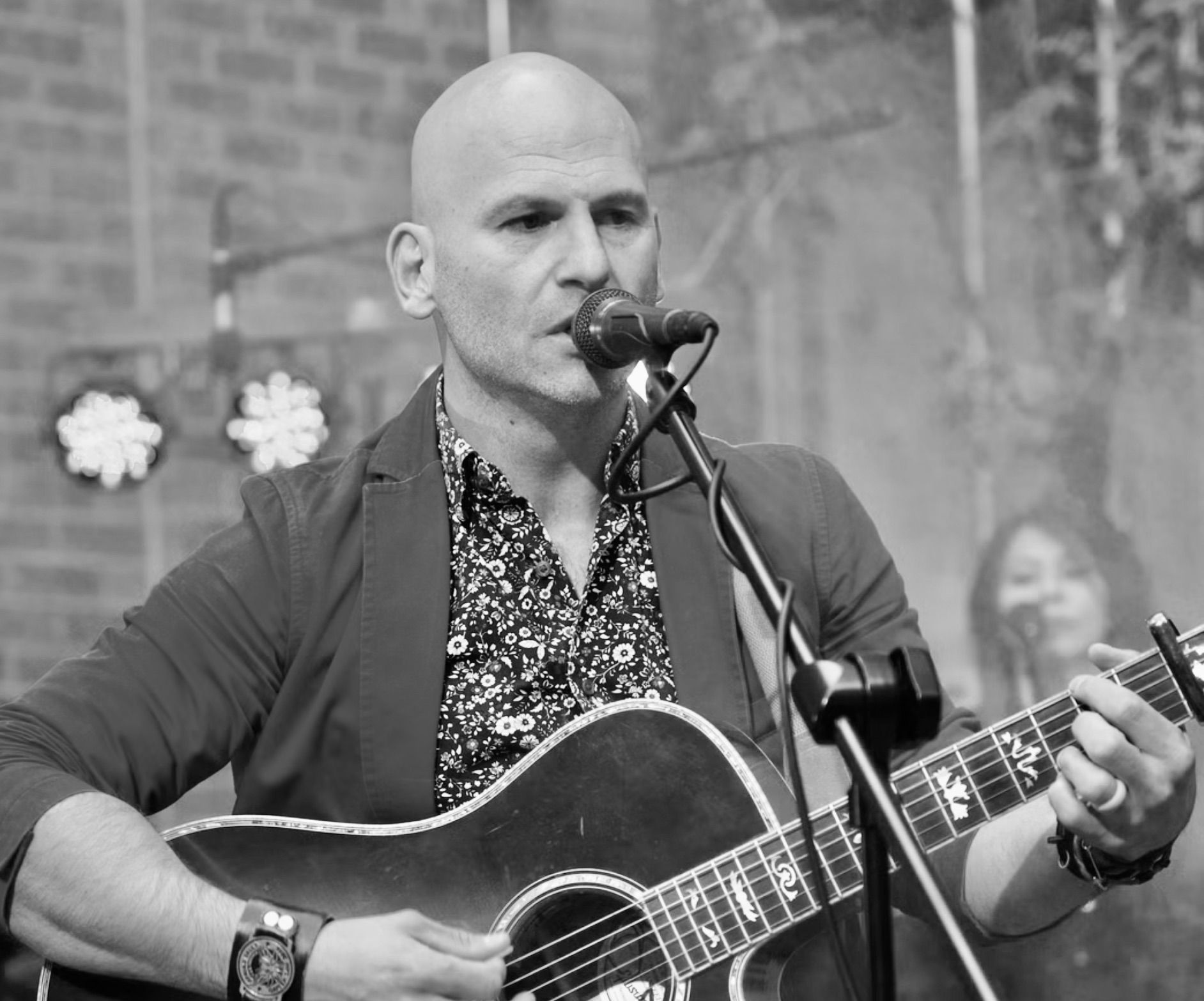 Bald man singing and playing acoustic guitar on stage, black and white.