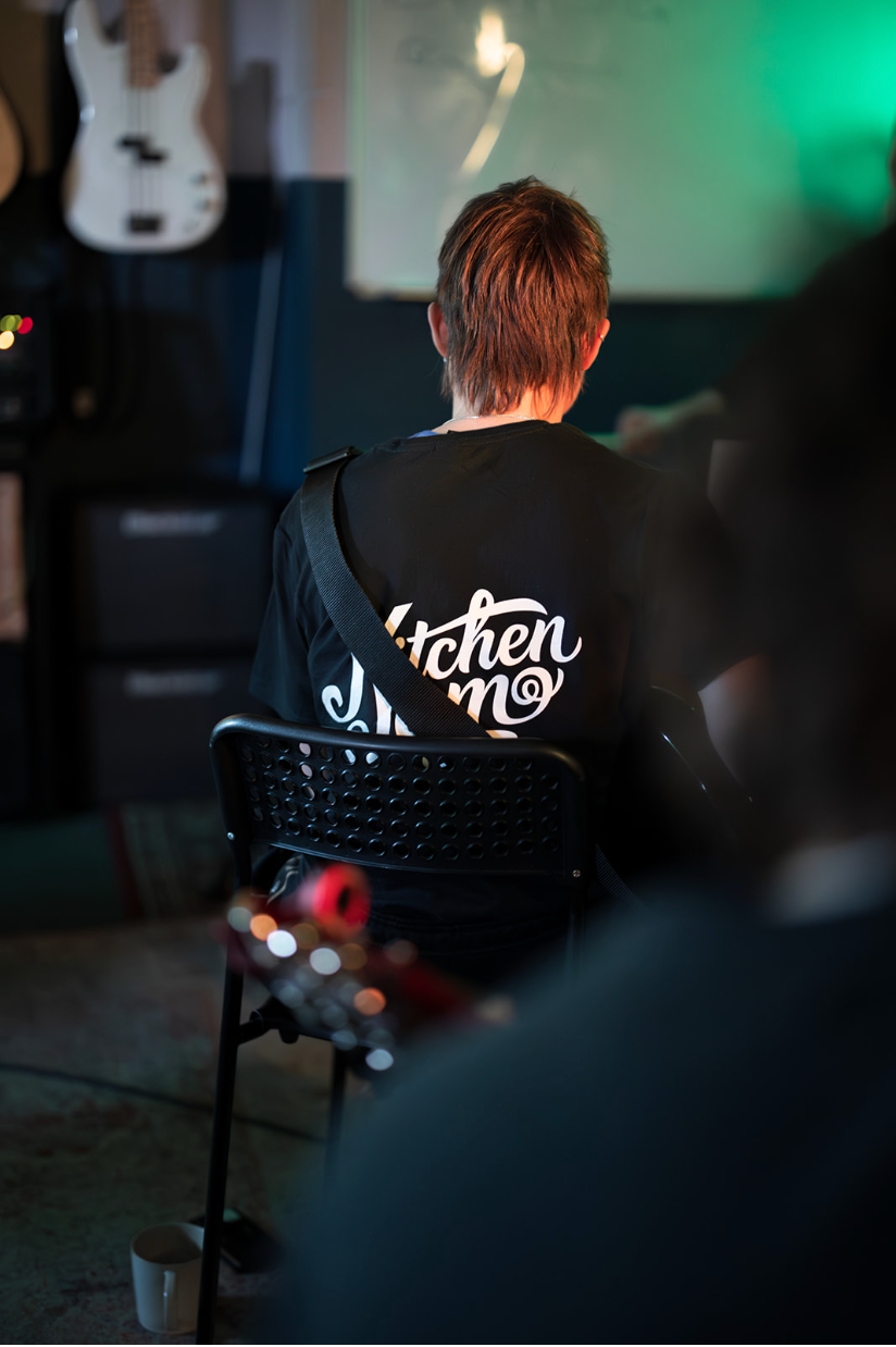 A man is sitting in a chair wearing a black shirt with the KitchenJam logo on it.