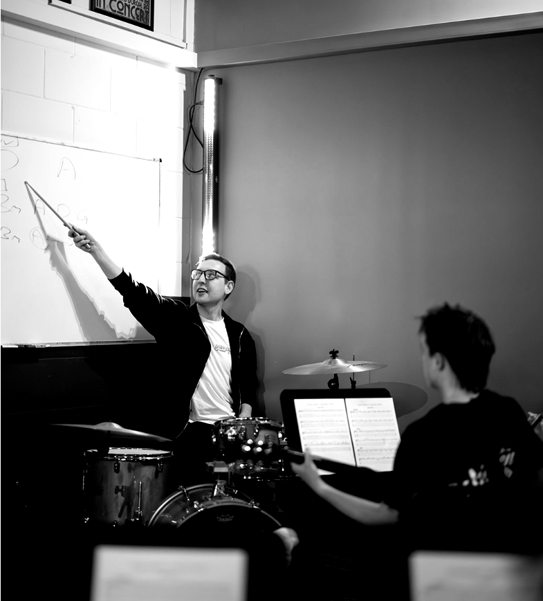 Man teaching music, pointing at whiteboard, with student, drums in room.